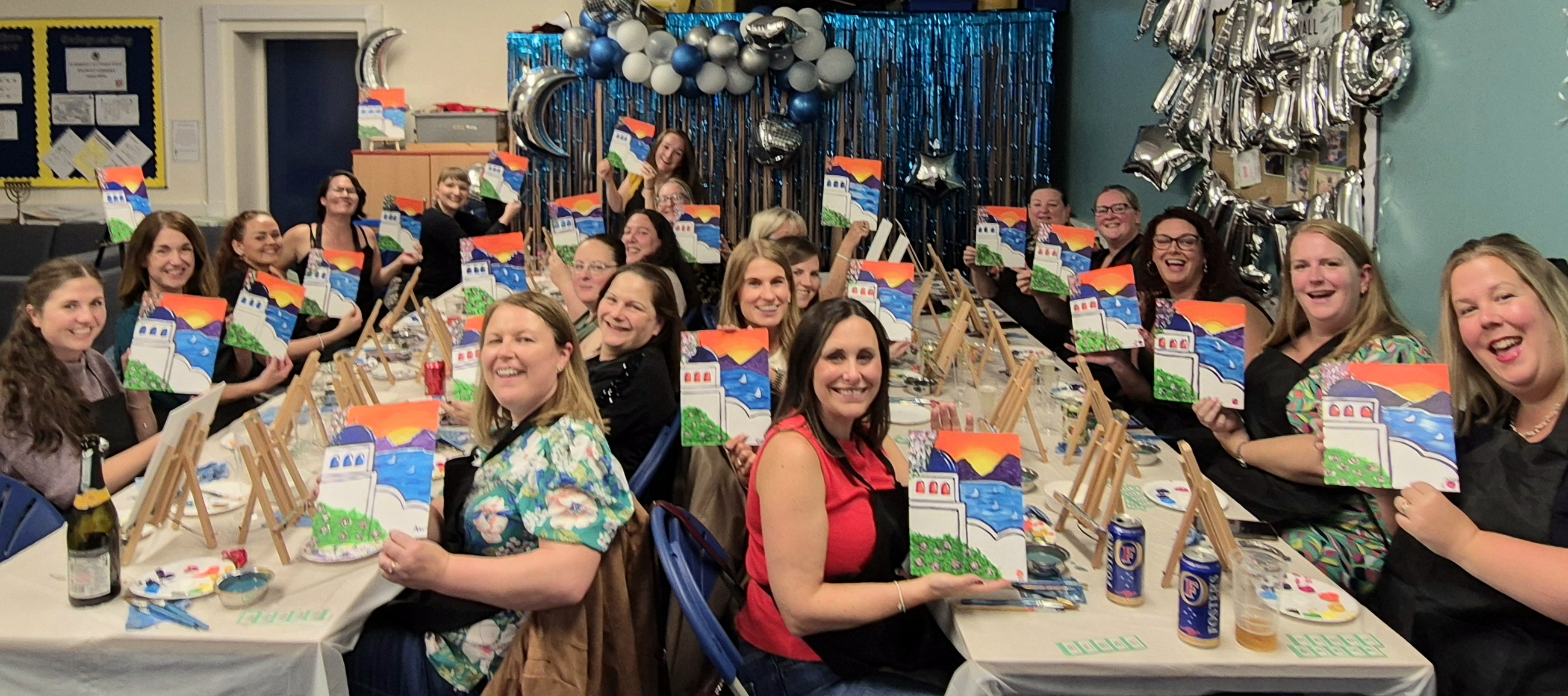 Photo of a large group of people seated at two long tables.  They are all holding up paintings.